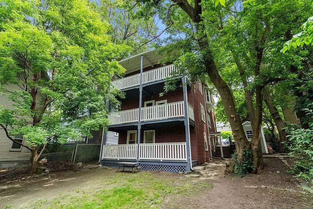 The back of a multi-story residential building with multiple balconies and a backyard surrounded by lush trees.