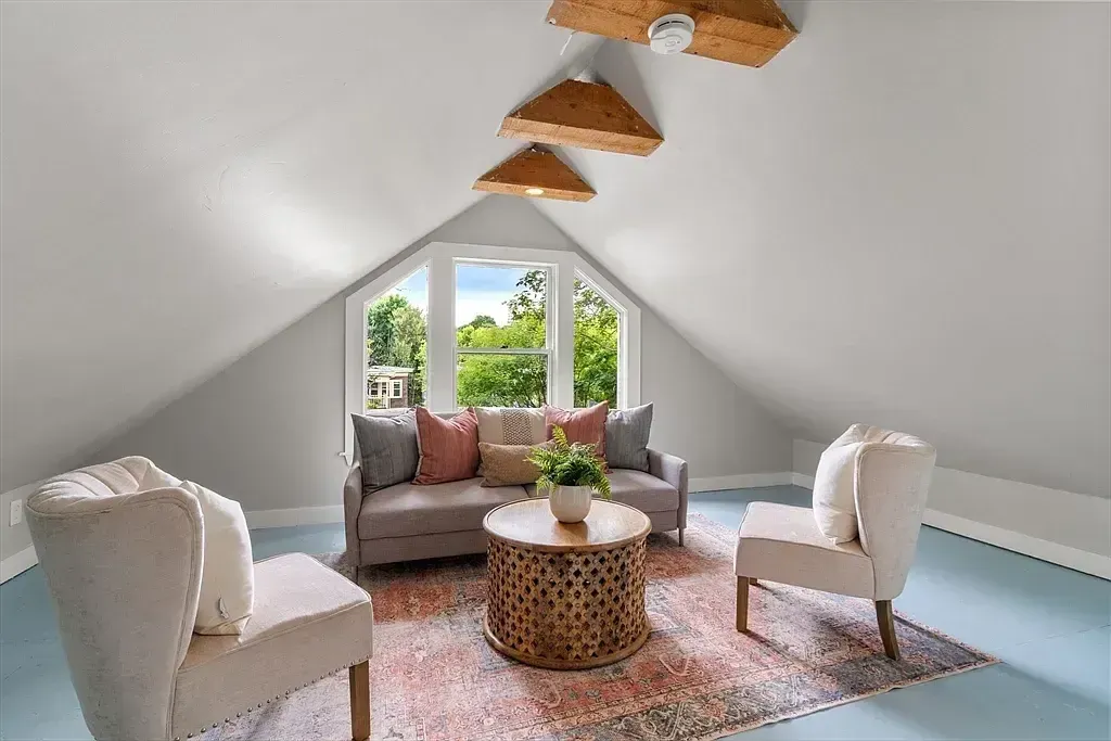 An attic sitting room features a sofa, two armchairs, a coffee table, a bay window, and slanted ceilings with exposed wooden beams.