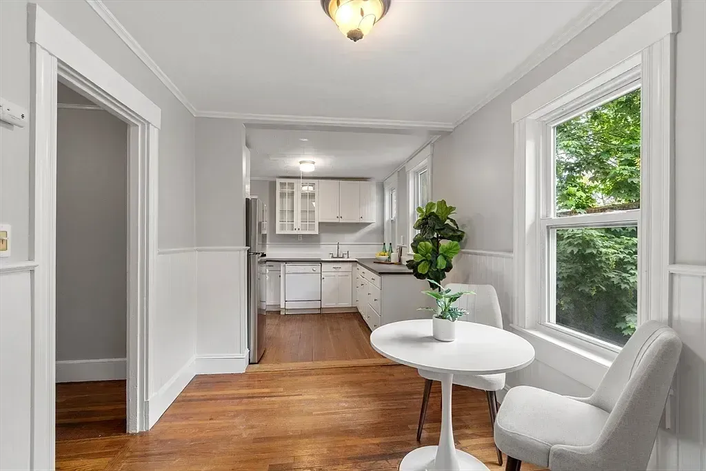 A dining area with a small white table and chairs is adjacent to a kitchen featuring white cabinets, a refrigerator, and a sink.