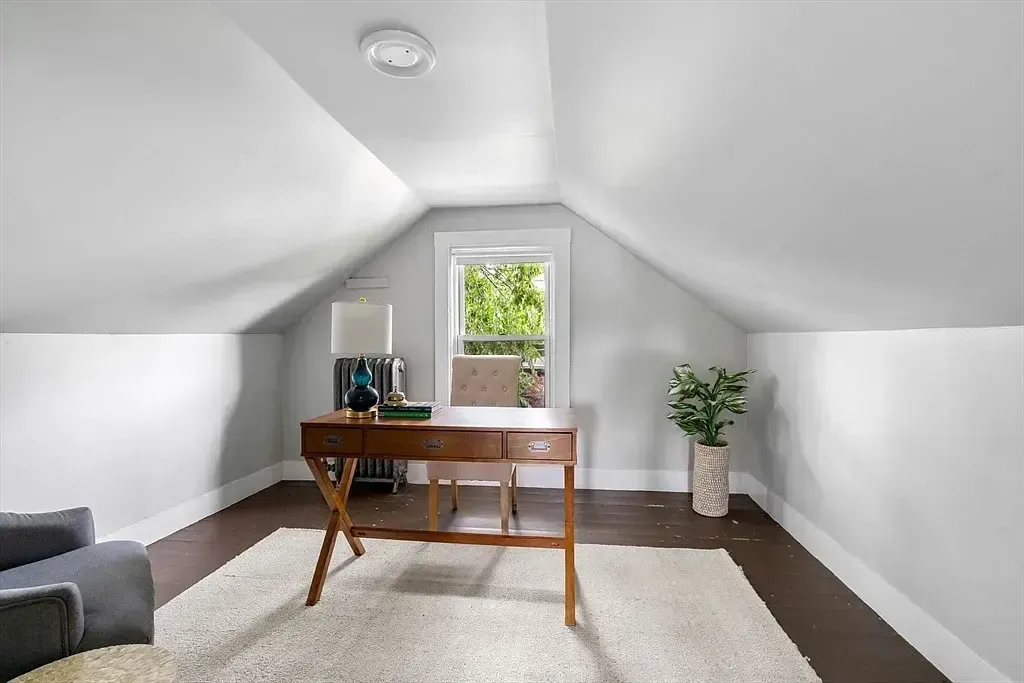 A home office in an attic space features a wooden desk, a chair, and a window overlooking trees.