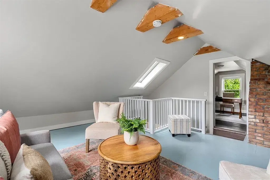 An attic living room features a skylight, exposed wooden beams, and a partial brick wall, with a railing separating a lower area.